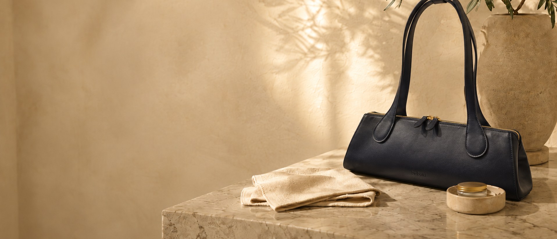 The.KDD in black resting on a marble ledge beside a soft cleaning cloth and a small balm, against a cream wall dappled with olive-branch shadows
