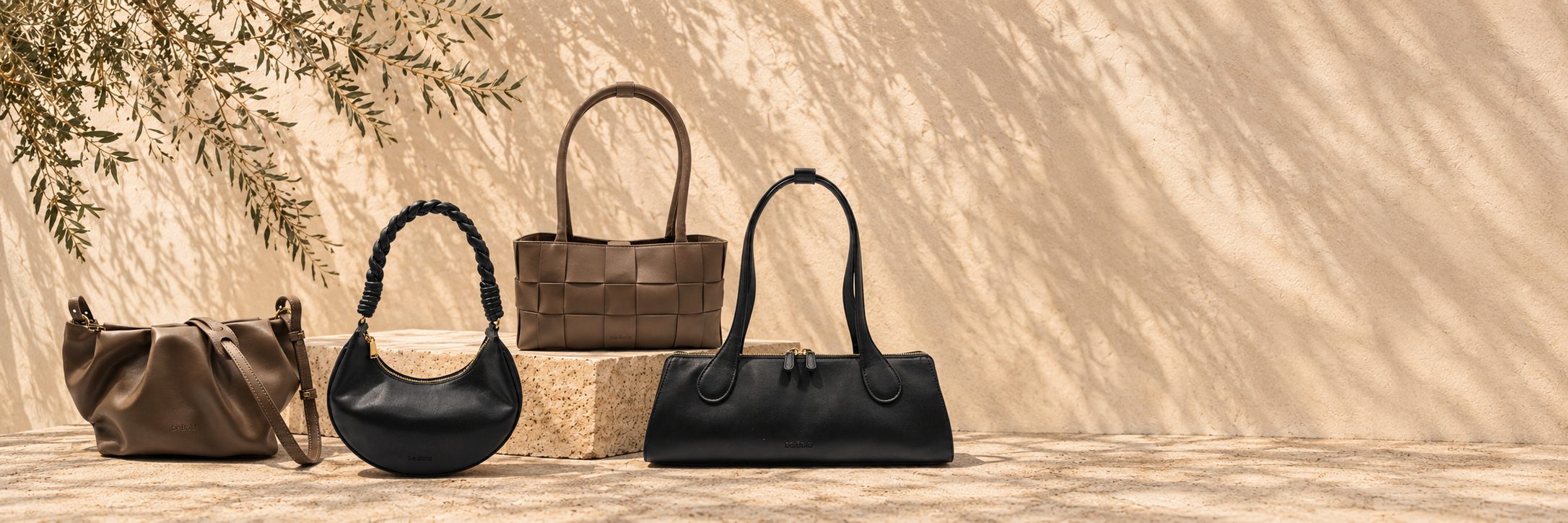 Four be.Bold handbags on a travertine ledge against a sunlit plaster wall, olive branch shadows falling across it