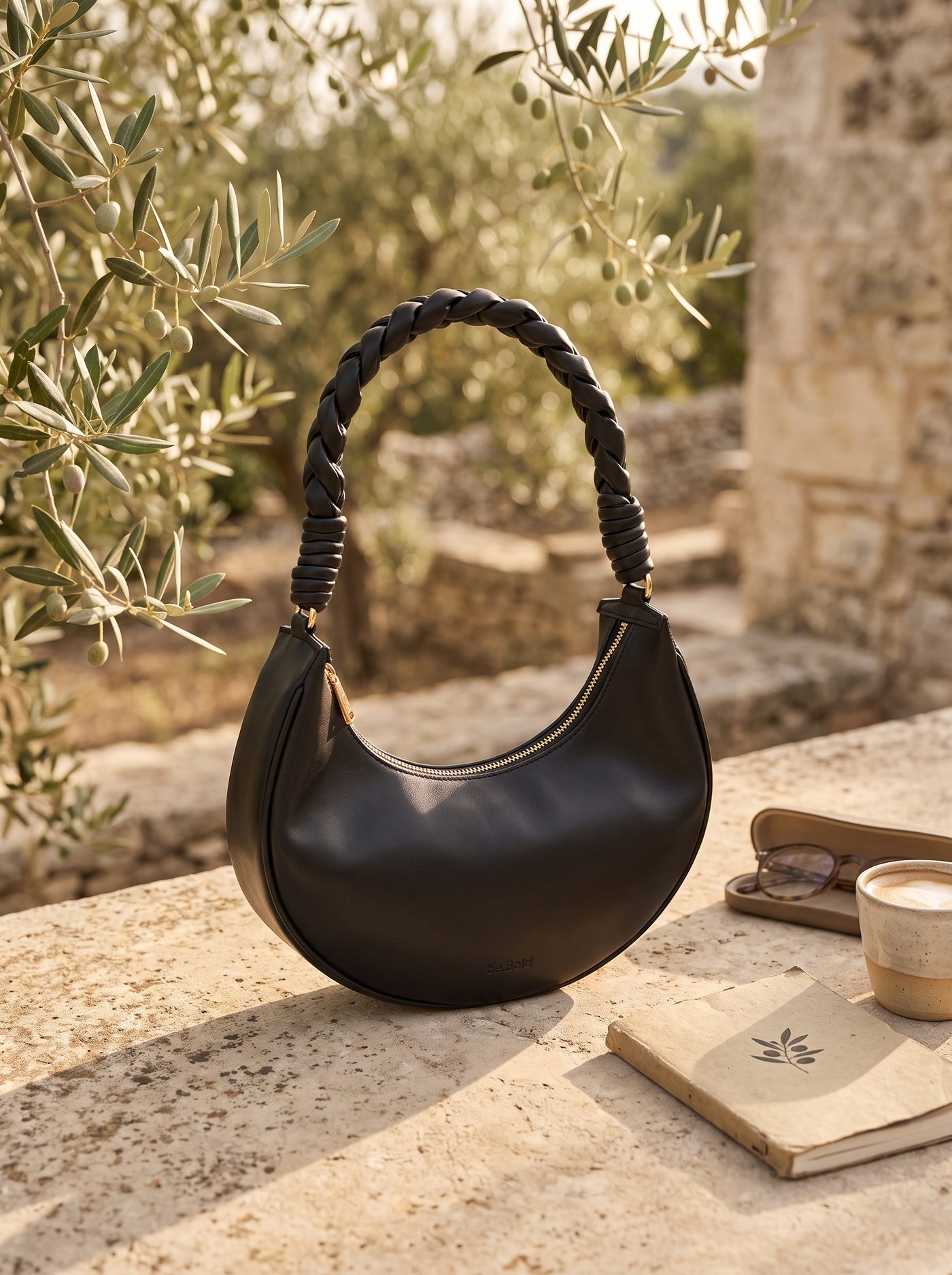 The.CP crescent shoulder bag in black Oleatex, its braided strap raised, on a travertine ledge beneath an olive tree